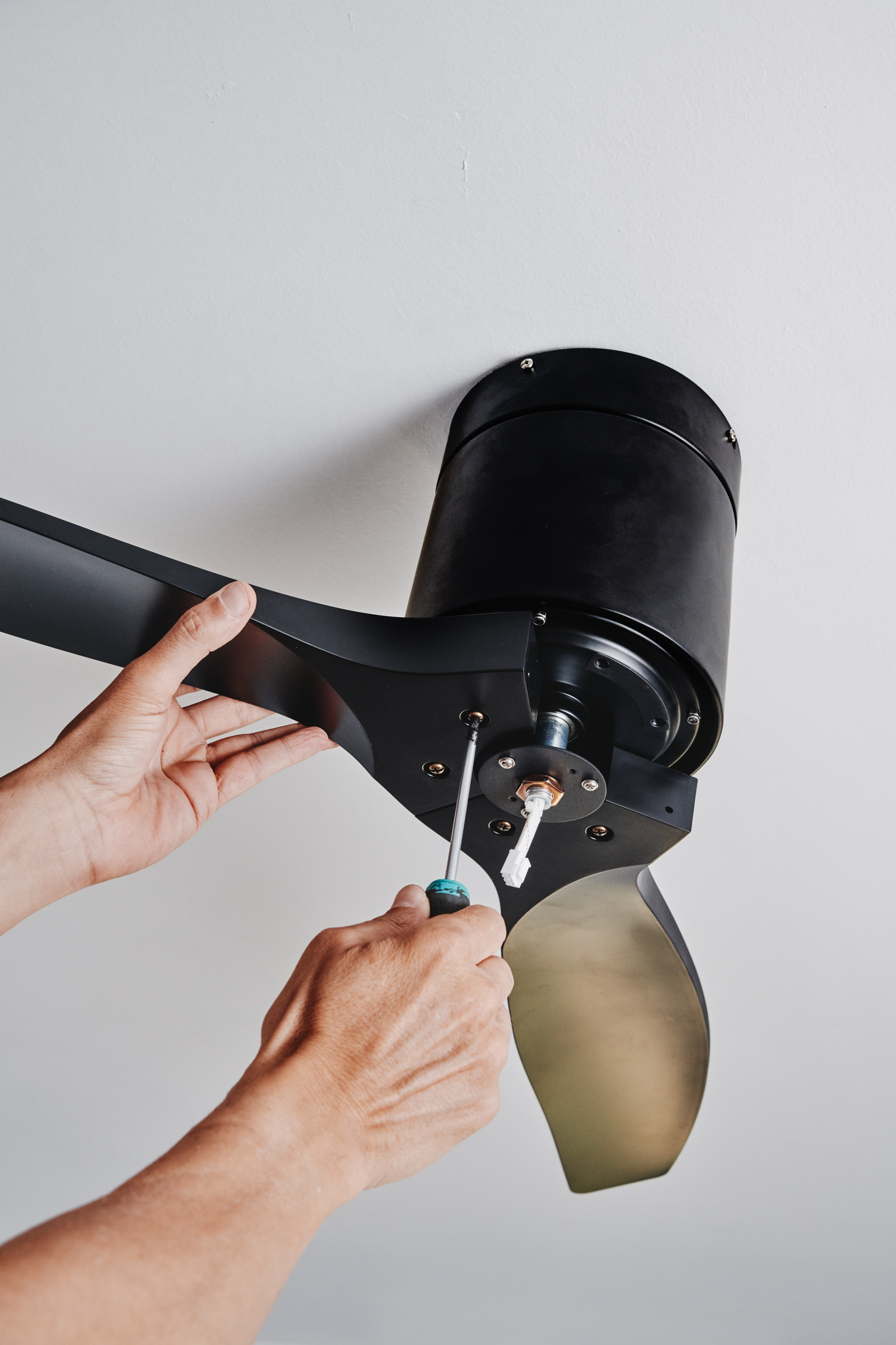 from directly below, a man secures the central housing of a black ceiling fan with a screwdriver as he steadies one curved blade during installation, with blank space above